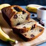Freshly baked banana bread with chunks of melted chocolate, sliced on a wooden cutting board. Ripe bananas and a cup of hot chocolate sit in the background, creating a cozy and inviting scene.