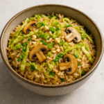 Close-up of a bowl filled with glossy jasmine rice tossed with soy sauce, thin ribbons of wilted Chinese cabbage, and sautéed mushroom slices, topped with chopped spring onions and cashew pieces, all presented in warm natural light against a neutral tabletop.