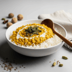 Professional food photography of a creamy yellow lentil dal served over white basmati rice in a white ceramic bowl, garnished with dried mint leaves and a drizzle of white cream or yogurt. The dish is photographed from above with a wooden spoon placed alongside, scattered whole spices including cardamom pods and cumin seeds around the bowl, and soft natural lighting against a neutral gray surface with a white linen napkin in the background.Retry