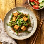 A ceramic plate with Filipino-inspired Smoky Adobong Sitaw featuring vibrant green beans and crispy golden tofu cubes atop jasmine rice, garnished with spring onions, and surrounded by fresh tomato and cucumber salad; styled on a rustic wooden table, softly lit, with a folded linen napkin and chopsticks as minimal props.