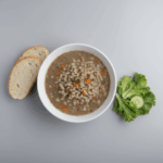 A stark, professional photograph of German-style barley soup in a plain white bowl, set against a neutral background. Beside the soup are two slices of plain crusty bread and a small bowl with a simple green salad, all under bright, clinical lighting to create a clean and boring aesthetic.