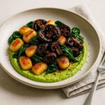 Crispy golden-brown air-fried gnocchi on a shallow ceramic plate with a swoosh of bright green lemon-pea cashew cream, topped with glossy miso-glazed charred mushrooms and wilted spinach, styled on a neutral stone background with a fork and folded napkin.