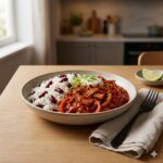 A close-up, high-angle shot of a ceramic bowl containing vegan Jackfruit Ropa Vieja. The dish features tender, shredded jackfruit simmered in a glossy, rich red tomato and pepper sauce on the right, served alongside "Congri"—white rice mixed with red kidney beans—on the left. The meal is garnished with a dollop of white vegan sour cream and fresh, sliced green spring onions. A beige linen napkin and black fork rest on the light wooden table beside the bowl, with a soft, blurred kitchen background.