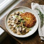 A top-down, 45-degree angle shot of a rustic, speckled ceramic plate on a dark, weathered wooden table. The plate features a golden-brown, crispy potato rösti with lacy edges and a creamy mushroom and white bean ragout. The ragout contains sliced brown champignons and king oyster mushrooms in a velvety, pale sauce, garnished with fresh thyme and scattered green spring onion rings. To the right of the plate, a folded off-white linen napkin sits with a small sprig of fresh thyme. Soft, natural light from the left creates gentle shadows, highlighting the textures of the food in a clean, editorial style.