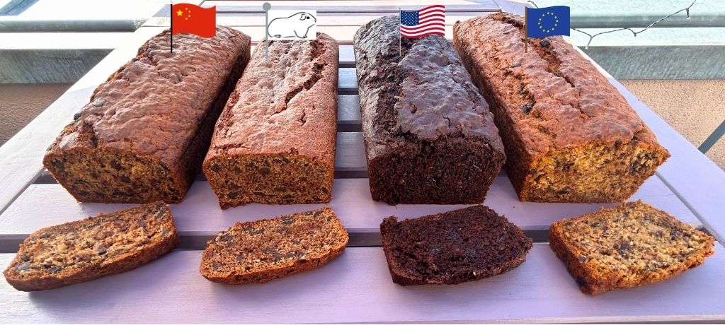 Four vegan banana bread loaves lined up side by side on a wooden terrace table, each marked with a small flag on a toothpick: Chinese flag for Qwen (far left, deep brown with chocolate chips), a white guinea pig flag for the human recipe (second from left, golden brown and compact), US flag for Gemini (second from right, very dark almost black from cocoa), and EU flag for Mistral (far right, golden and evenly risen). In front of each loaf lies a single cut slice, clearly showing the distinct colour and texture differences between the four recipes. Bright outdoor daylight.