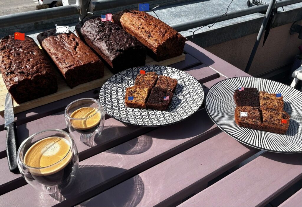 Four freshly baked vegan banana bread loaves arranged on a wooden cutting board on an outdoor terrace table, each labelled with a small flag: Chinese flag for Qwen, US flag for Gemini, EU flag for Mistral, and a white guinea pig flag for the human recipe. In the foreground, two patterned plates hold slices of all four breads side by side for tasting, each slice also marked with its corresponding flag. Two double-walled glass espresso cups sit beside the plates in the sunshine.
