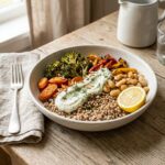 A styled editorial food photograph in square format. A wide, shallow off-white speckled stoneware bowl on a warm oak wooden surface holds the Baltic Bowl: a base of earthy buckwheat topped with roasted broccoli florets, golden-edged carrot rounds, charred yellow and red bell pepper strips, and plump white beans. A swoosh of pale creamy dill cashew sauce runs across the centre, garnished with fresh dill and toasted sunflower seeds. A halved lemon sits on the rim. A folded beige linen napkin and matte silver fork rest to the left. Soft natural light enters from the upper left, casting gentle warm shadows. Shallow depth of field blurs the white ceramic jug in the background.