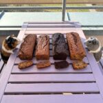 A bright, outdoor photo of four different freshly baked banana bread loaves lined up on a light purple slatted wooden table on a sunny balcony. Each loaf has a matching slice cut and placed directly in front of it to show the interior texture. From left to right: a dark brown loaf with chocolate chips, a compact golden-brown loaf, a very dark chocolate loaf, and a tall, golden loaf. At opposite ends of the lineup, two realistic, fluffy guinea pigs rest their front paws on the table, looking curiously toward the freshly baked bread.