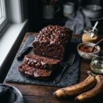 A professional, moody food photograph of a deeply rich, sliced vegan double chocolate banana bread resting on a dark slate board. The loaf is dark brown from cocoa powder, generously studded throughout and topped with glossy, melting chocolate chips. Surrounding the bread are key recipe ingredients: a rustic bowl of cocoa powder, a jar of raw cane sugar, a small pitcher of melted coconut oil, and a few speckled overripe bananas. The lighting is decadent and atmospheric, highlighting the melted chocolate pools and the moist texture of the crumb.