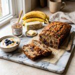 A professional, high-resolution food photograph of a freshly baked, sliced vegan banana bread on a white marble cutting board. The loaf has a beautifully risen, golden-brown crust and a fluffy interior with visible chocolate chips. Around the board, key ingredients from the recipe are artfully arranged: a small glass bowl of creamy vegan yogurt, a jar of ground flaxseed, a pat of vegan butter, and ripe, speckled bananas. The lighting is soft and natural, creating an inviting, elegant bakery aesthetic.