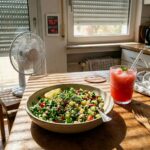 Square editorial food photo of a chilled bowl of vegan millet tabbouleh with parsley, cucumber, tomato, and lemon, served beside a frosty watermelon slush in a hot German apartment kitchen with closed blinds, a closed window, a small fan, and a digital display reading 38.3°C and 45% humidity.